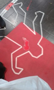 Nachgestellter Tatort eines Mordes: Körperumriss, der mit Klebeband auf einen Fußboden angedeutet wurde. Ein roter Fleck suggeriert Blut.
